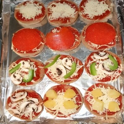 Twelve mini pizzas on a foil-lined tray, topped with tomato sauce, cheese, green peppers, mushrooms, and pineapple—these kid friendly go to meals offer fun and tasty combinations for everyone to enjoy.