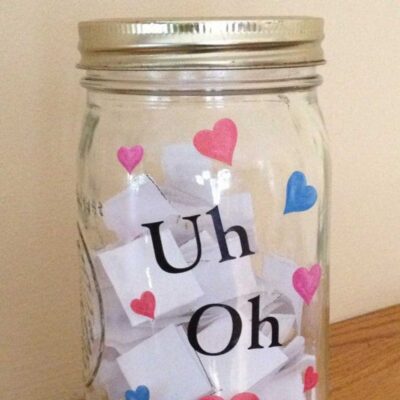 A glass consequence jar with a metal lid labeled "Uh Oh," decorated with pink and blue hearts, containing folded slips of white paper inside.