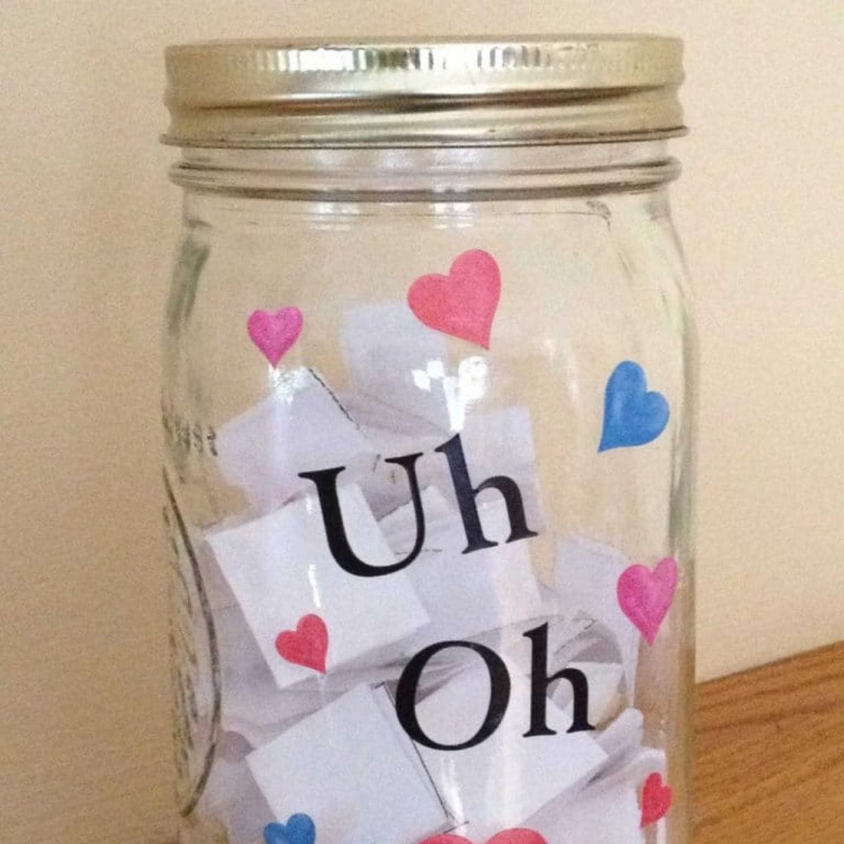 A glass consequence jar with a metal lid labeled "Uh Oh," decorated with pink and blue hearts, containing folded slips of white paper inside.