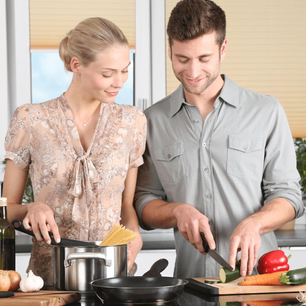 A woman adds spaghetti to a pot while a man slices a cucumber on a cutting board in a modern kitchen, making meal prep for easy dinner parties with fresh vegetables and cookware.