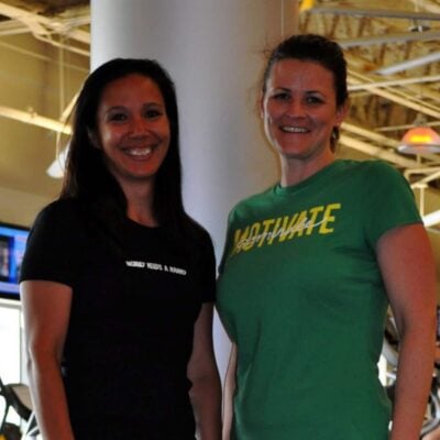 Two women stand side by side in a gym, both smiling. One wears a black shirt, the other a green shirt with "MOTIVATE" on it—ready to tackle a fast full body workout. Gym equipment can be seen in the background.