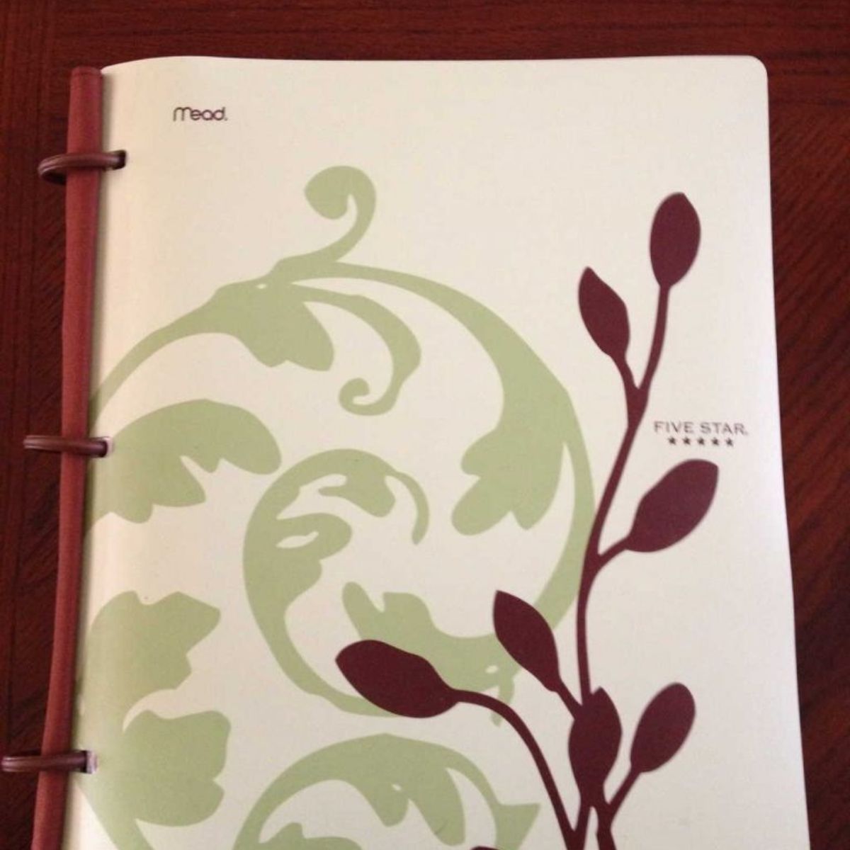 A Mead Five Star notebook with a white cover featuring green and maroon plant designs, perfect as a stylish family binder, placed on a wooden surface.