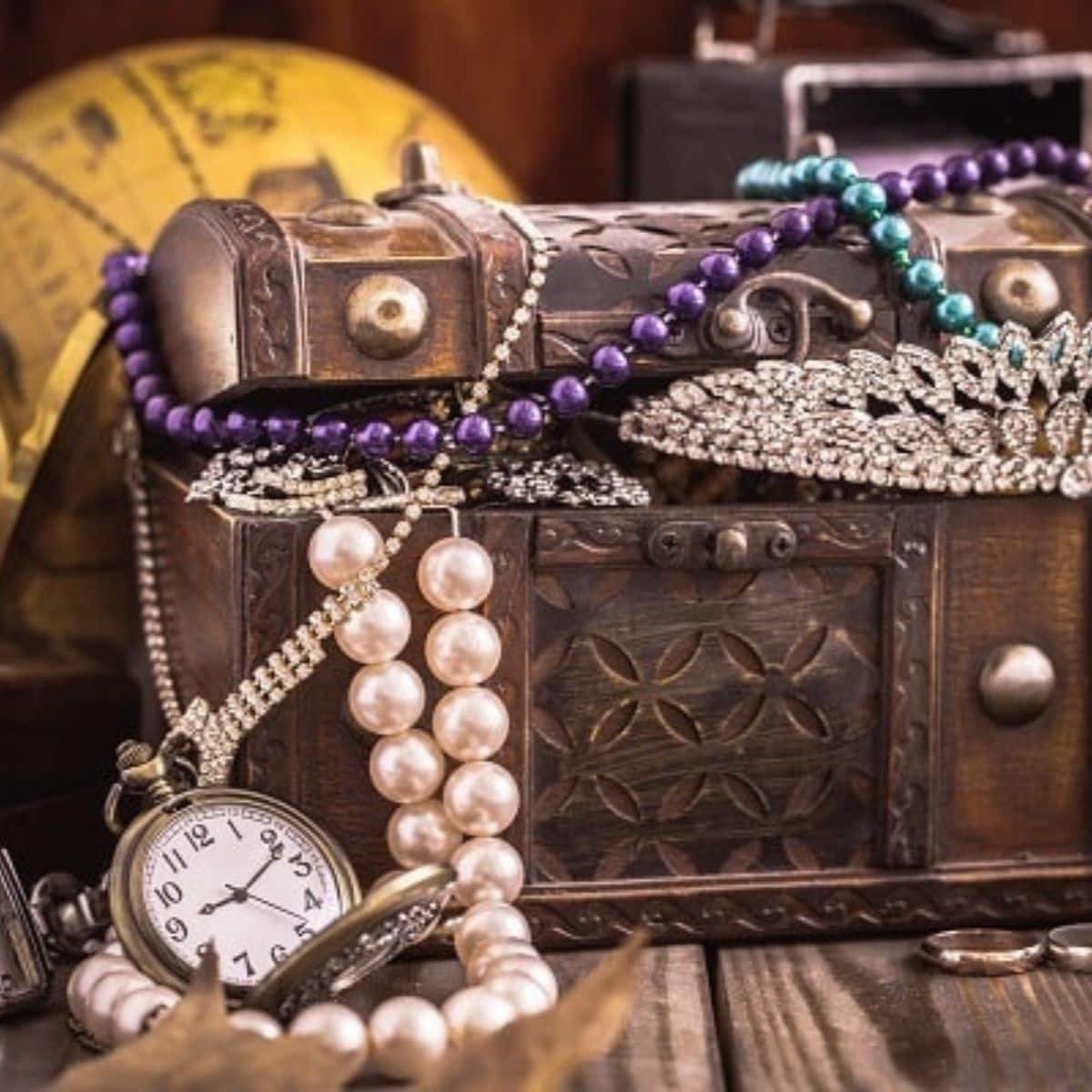 An ornate treasure chest, perfect for treasure hunts, is filled with pearl necklaces, beaded jewelry, a jeweled tiara, and a pocket watch resting on a wooden surface.