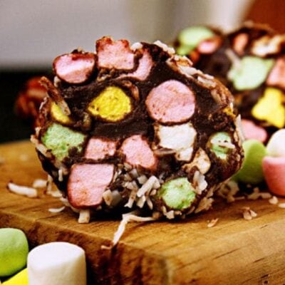 Close-up of a slice of chocolate dessert inspired by the cathedral windows recipe, topped with colorful mini marshmallows and shredded coconut on a wooden surface.