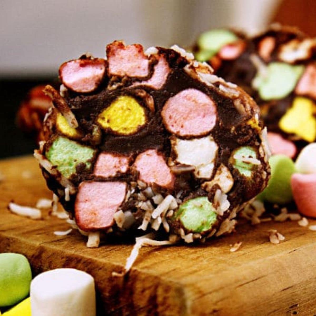 Close-up of a slice of chocolate dessert inspired by the cathedral windows recipe, topped with colorful mini marshmallows and shredded coconut on a wooden surface.