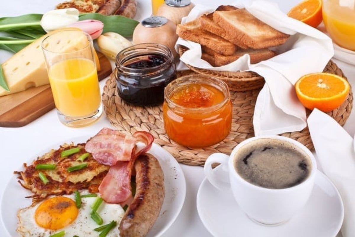 Breakfast spread with fried eggs, bacon, sausage, toast, sliced cheese, orange juice, coffee, orange slices, and jars of jam on a table.