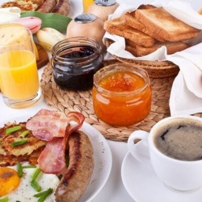 A breakfast spread with fried eggs, bacon, sausage, toast, orange juice, coffee, and jars of jam arranged on a table.
