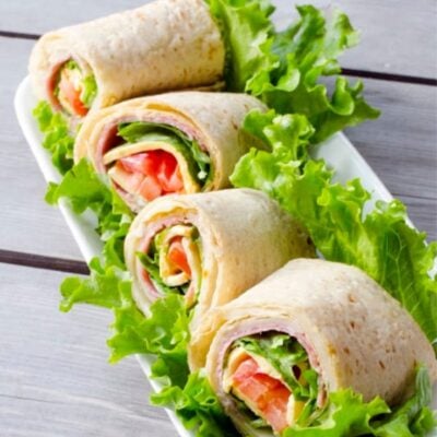 A rectangular white plate with four tortilla wraps filled with lettuce, tomato, and other deli ingredients, garnished with loose lettuce leaves.