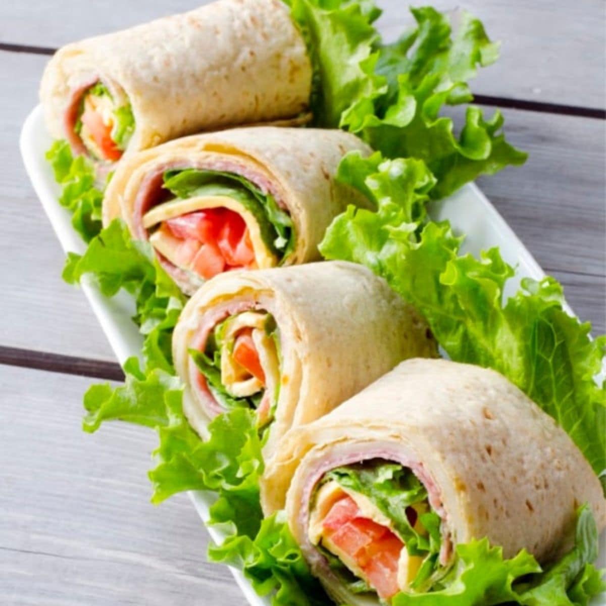 A rectangular white plate with four tortilla wraps filled with lettuce, tomato, and other deli ingredients, garnished with loose lettuce leaves.