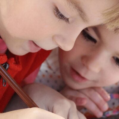 Two children closely look at something, with one holding a pencil and the other observing attentively—a great way to keep your kids busy at a restaurant.