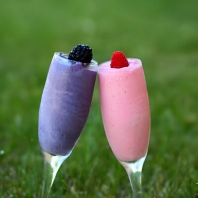 Two champagne glasses filled with purple and pink frozen drinks, each topped with a berry, are tilted toward each other on a grassy background—perfect inspiration for easy healthy smoothie recipes.