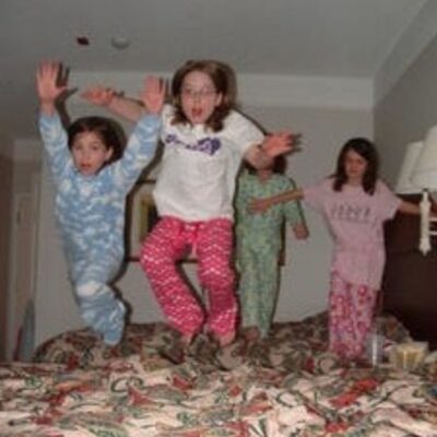 Four children in pajamas are jumping on a bed in a hotel room, creating a lively, stress free slumber party—two soar in the foreground while two more are ready to jump in the background.