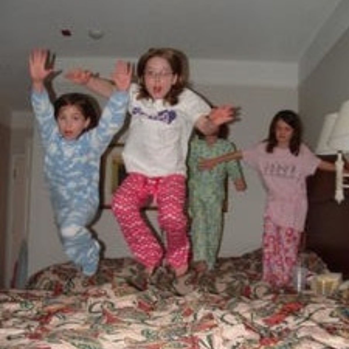 Four children in pajamas are jumping on a bed in a hotel room, creating a lively, stress free slumber party—two soar in the foreground while two more are ready to jump in the background.