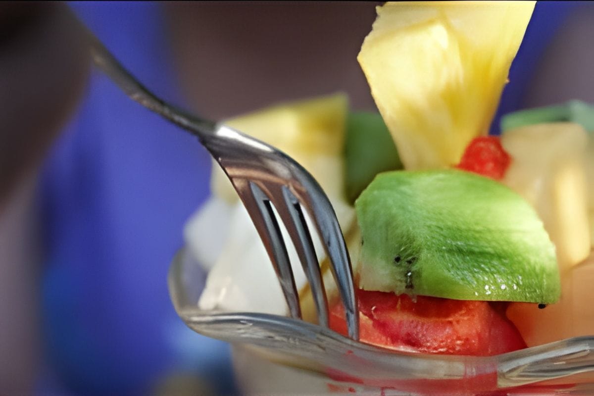 A close-up of a fork picking up a piece of kiwi from a glass bowl filled with assorted fresh fruit, showing how to eat healthy for life with vibrant options like pineapple, watermelon, and melon.