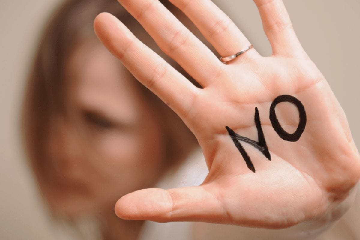 A person demonstrates how to say no by holding up their hand with the word "NO" written on the palm, their face blurred in the background.