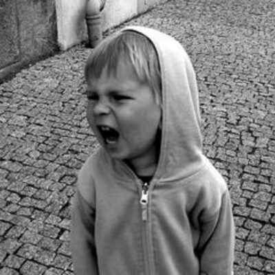 A young child in a hooded jacket stands on a cobblestone street, mouth open wide as if shouting—a moment that highlights the importance of practicing no yell discipline.