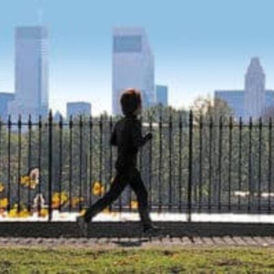 Person jogging on a paved path near a black metal fence with city skyscrapers and trees in the background—a great spot for running for beginners.