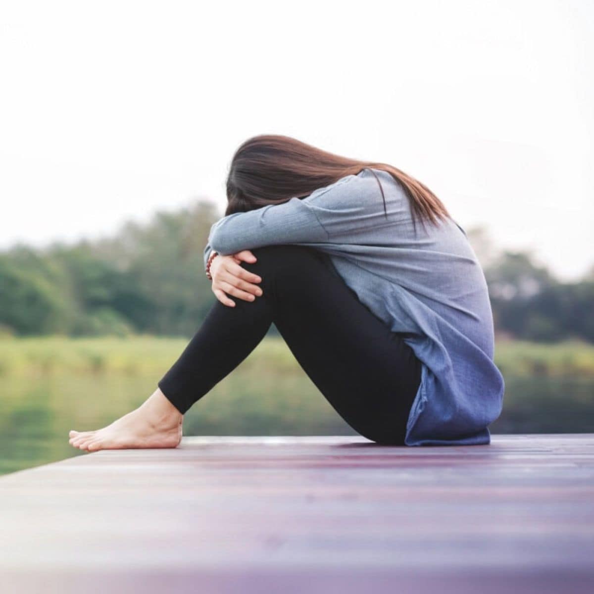 A person sits on a wooden dock by the water, hugging their knees with their head down, appearing contemplative or sad. Trees and water are in the background.