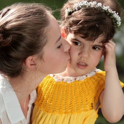 A woman kisses a young girl on the cheek, capturing a candid moment of bonding with your daughter. The girl, in a yellow dress and floral headband, looks away with a slightly uncomfortable expression.