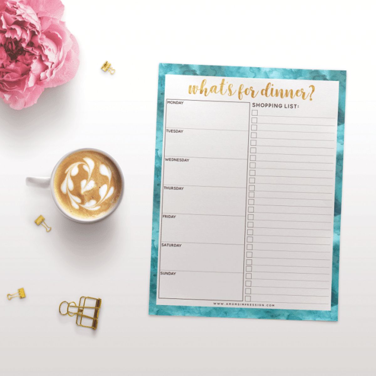 A weekly meal planning dinner planner with a shopping list, a cup of latte with latte art, a pink flower, and gold paper clips arranged on a white surface.