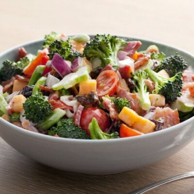 A bowl of salad featuring broccoli, cherry tomatoes, red onions, cheese cubes, bacon bits, and raisins sits on a wooden table—perfect for your Memorial Day menu.