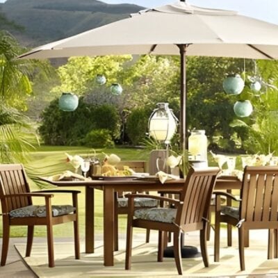 A wooden outdoor dining set with four chairs and a beige umbrella is arranged on a patio, surrounded by greenery and hanging lanterns, with hills visible in the background.