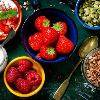 A bowl of strawberries surrounded by bowls of raspberries, granola, yogurt with strawberries, pumpkin seeds, a spoon, and a jar of milk on a green surface—perfect greek yogurt topping ideas for a vibrant breakfast spread.