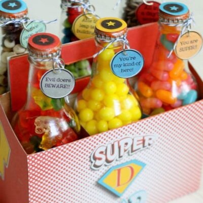Four glass bottles filled with colorful candy are arranged in a carrier labeled "SUPER." Each bottle has a tag with a superhero-themed message, making this one of the perfect DIY Father’s Day ideas for a fun and thoughtful gift.