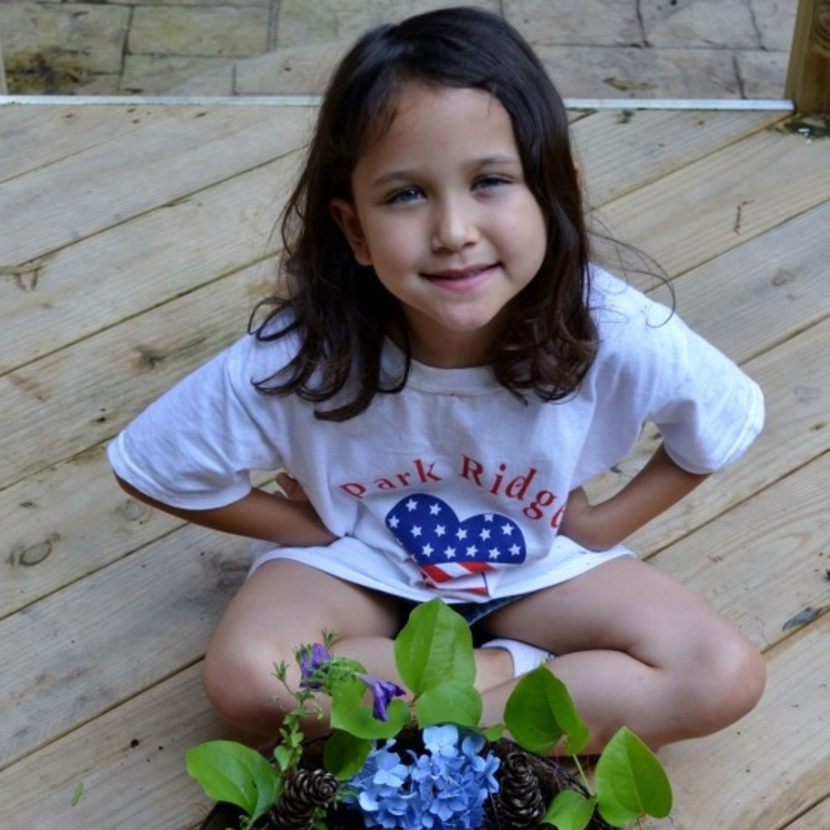Young girl with brown hair sits on a wooden deck, smiling at the camera. She is wearing a "Park Ridge" t-shirt and is next to a planter with green leaves, blue flowers, and a tiny fairy garden peeking through.