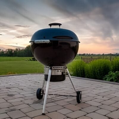 A black charcoal kettle grill with a lid stands on a paved patio, ready for your favorite grill recipe, with green grass and bushes in the background at sunset.