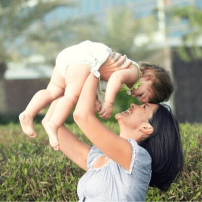 Woman holding a smiling baby above her head outdoors, both looking at each other, with greenery and blurred trees in the background—one of many easy ways to be active with your kids.