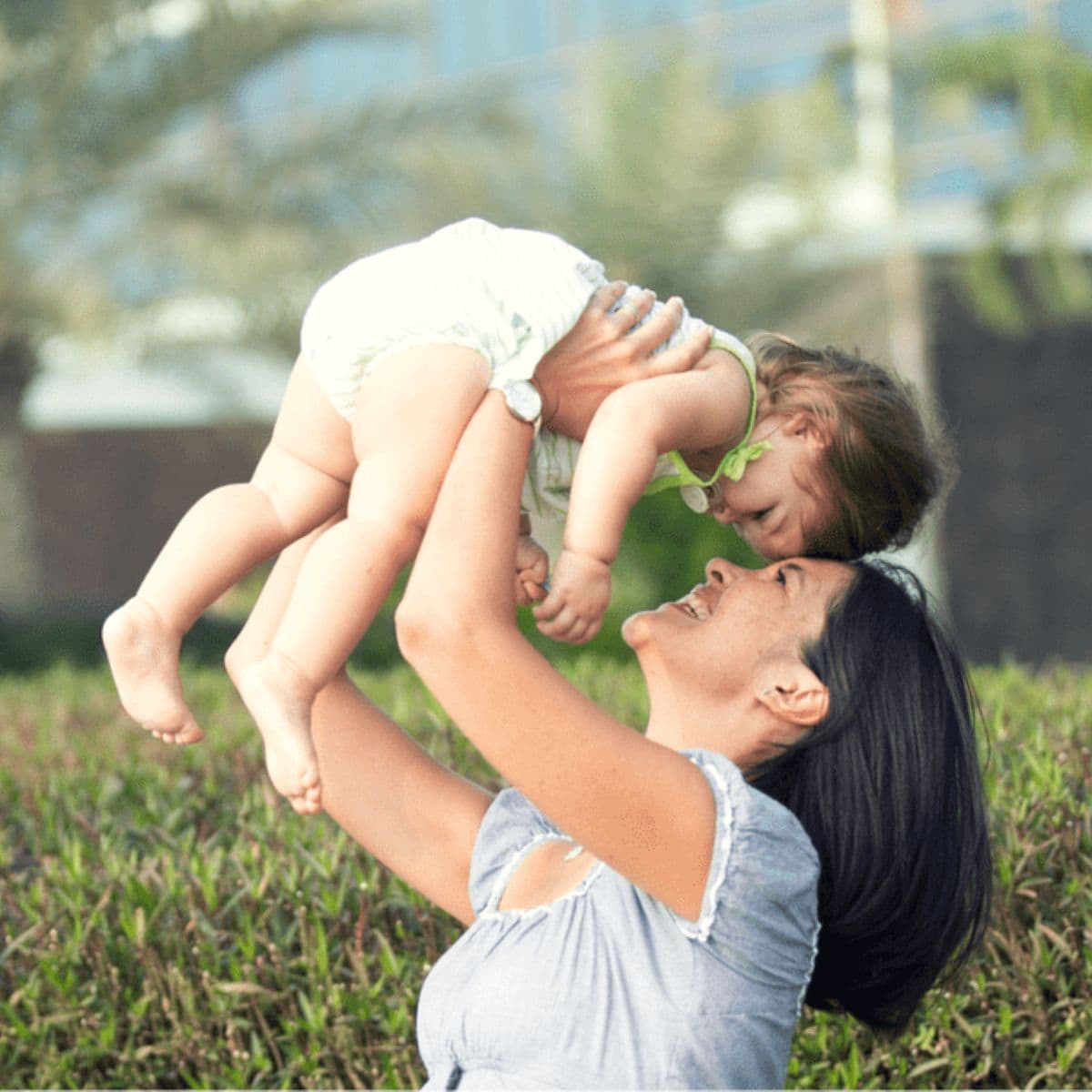 Woman holding a smiling baby above her head outdoors, both looking at each other, with greenery and blurred trees in the background—one of many easy ways to be active with your kids.