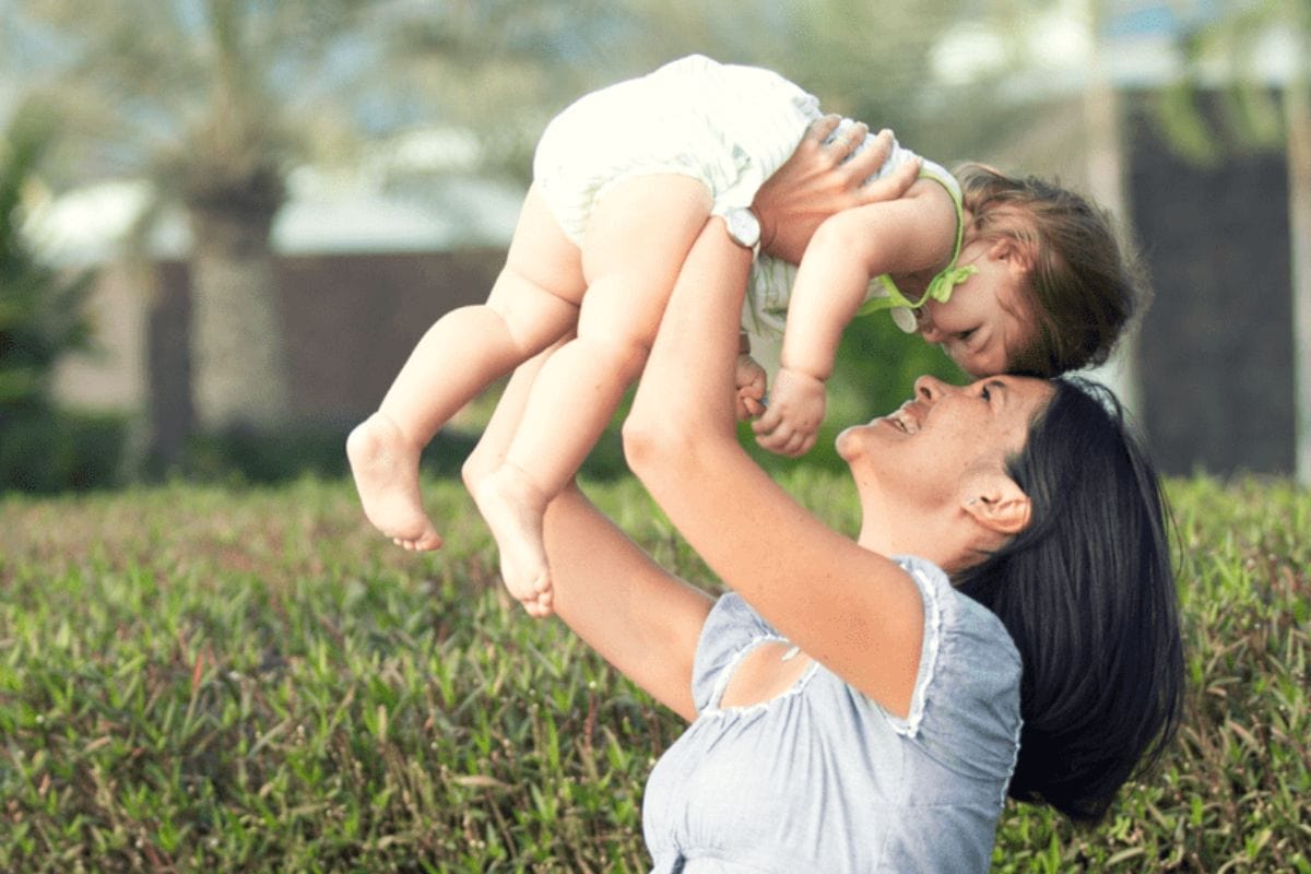 A woman outdoors lifts a smiling baby above her head, both making eye contact-one of many easy ways to be active with your kids. Green bushes and trees are visible in the background.