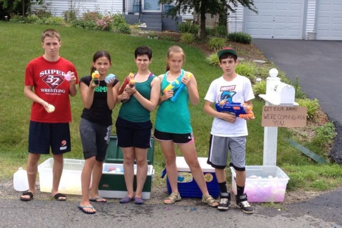 Five teenagers stand on a driveway holding water balloons and squirt guns, surrounded by tubs of water balloons and a sign reading "Get your ammo, we're coming for you"-showing easy ways to be active with your kids while having fun outdoors.