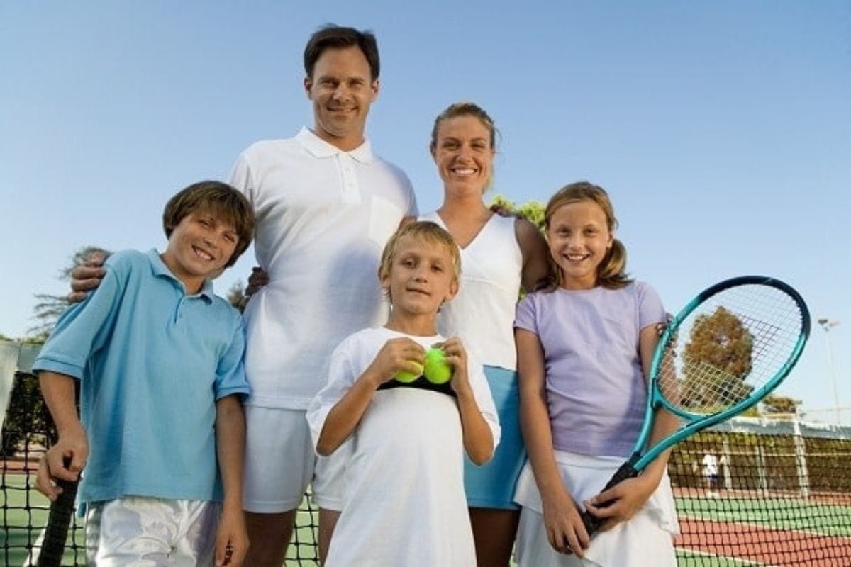 A family of five poses on a tennis court-two adults and three children, one holding tennis balls and another a racket-showcasing easy ways to be active with your kids.