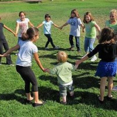 A group of children stand on grass holding hands in a circle, enjoying group activities for kids as they play together outdoors on a sunny day.