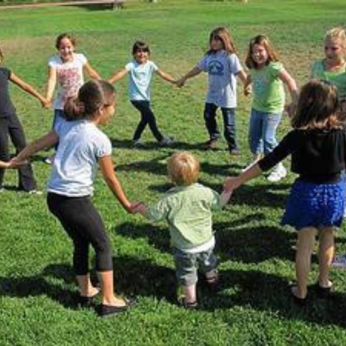 A group of children stand on grass holding hands in a circle, enjoying group activities for kids as they play together outdoors on a sunny day.