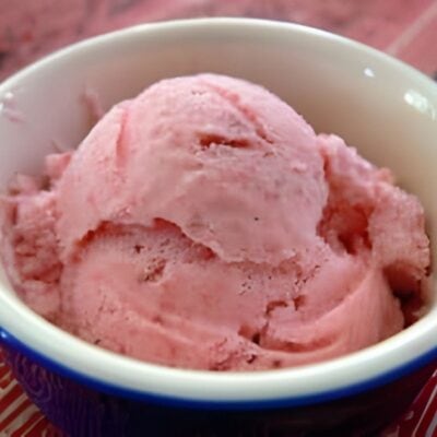 A scoop of pink strawberry ice cream in a white bowl with a blue rim, placed on a red surface—perfect for those seeking healthy desserts with vibrant flavors.