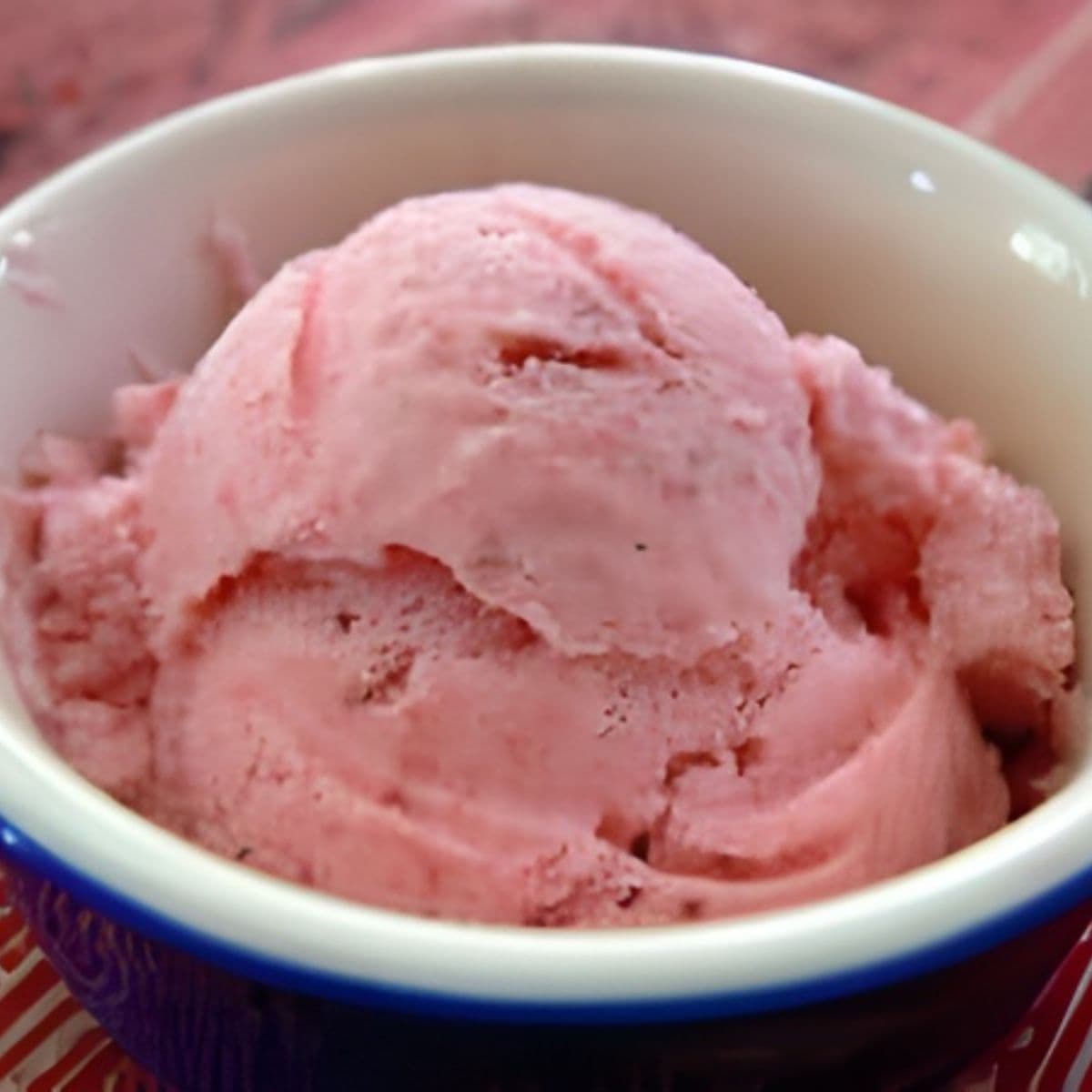 A scoop of pink strawberry ice cream in a white bowl with a blue rim, placed on a red surface—perfect for those seeking healthy desserts with vibrant flavors.