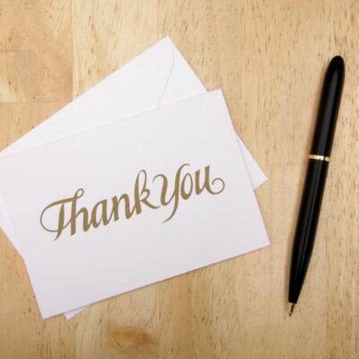 A thank you card with gold lettering sits on top of a white envelope next to a black pen on a wooden surface.