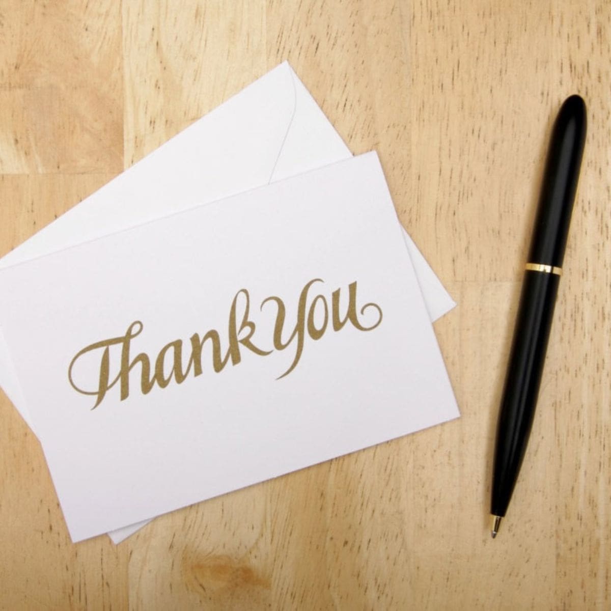 A thank you card with gold lettering sits on top of a white envelope next to a black pen on a wooden surface.