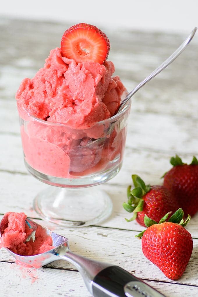 homemade strawberry frozen yogurt in a glass with a spoon in it on a wooden table next to strawberries and an ice cream scoop