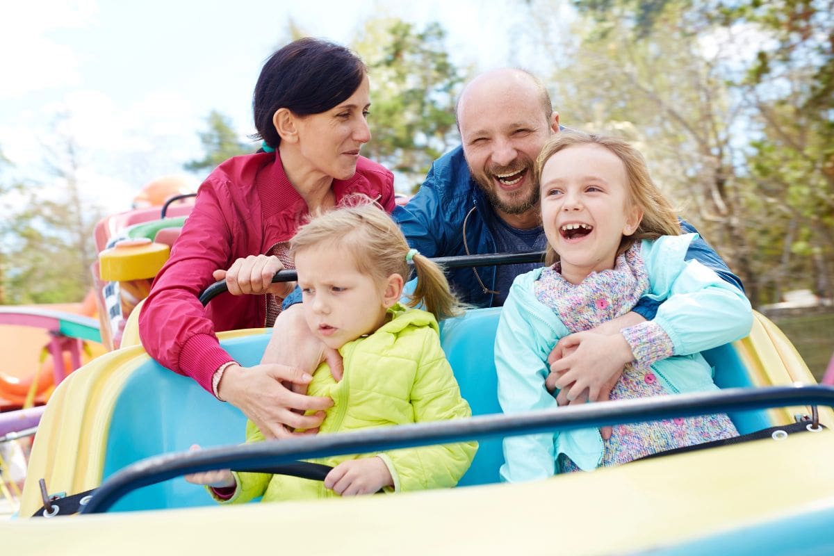 A family of four enjoys a colorful amusement park ride outdoors; two adults sit with two young girls, one smiling and the other serious.