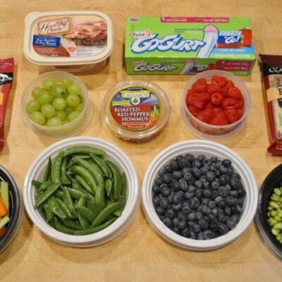 Assorted healthy snacks including grapes, raspberries, blueberries, snap peas, hummus, edamame, cheese sticks, yogurt tubes, and packaged deli meat arranged on a wooden surface.