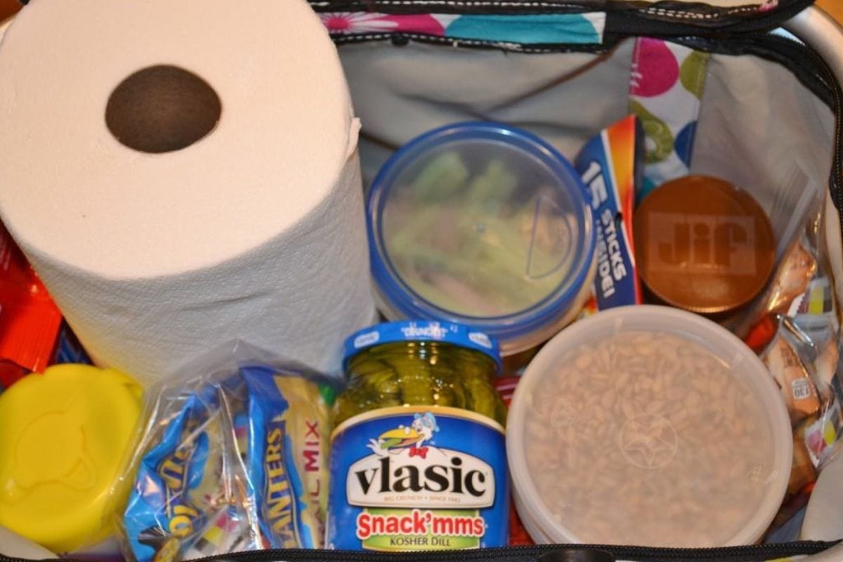 An open bag contains a roll of paper towels, jars of peanut butter and pickles, a plastic container, snack bars, and other assorted packaged food items.