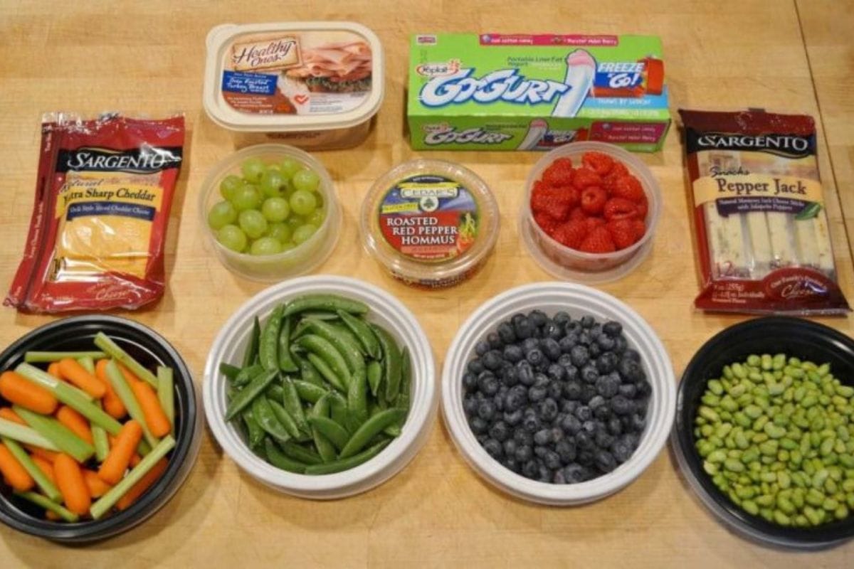 Assorted snacks including cheese, hummus, yogurt, grapes, raspberries, blueberries, snap peas, edamame, baby carrots, and celery arranged on a wooden surface.