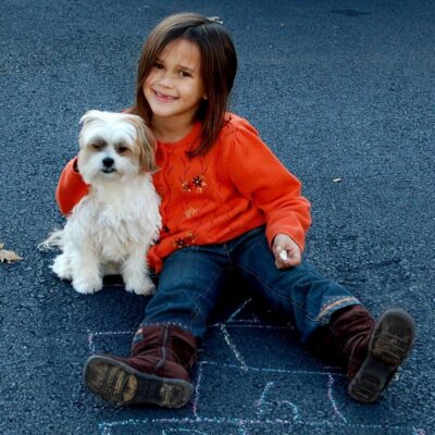 A young girl in an orange sweater sits on asphalt next to a small white dog, holding chalk with a hopscotch pattern drawn beneath them, demonstrating child safety with dogs during playtime.