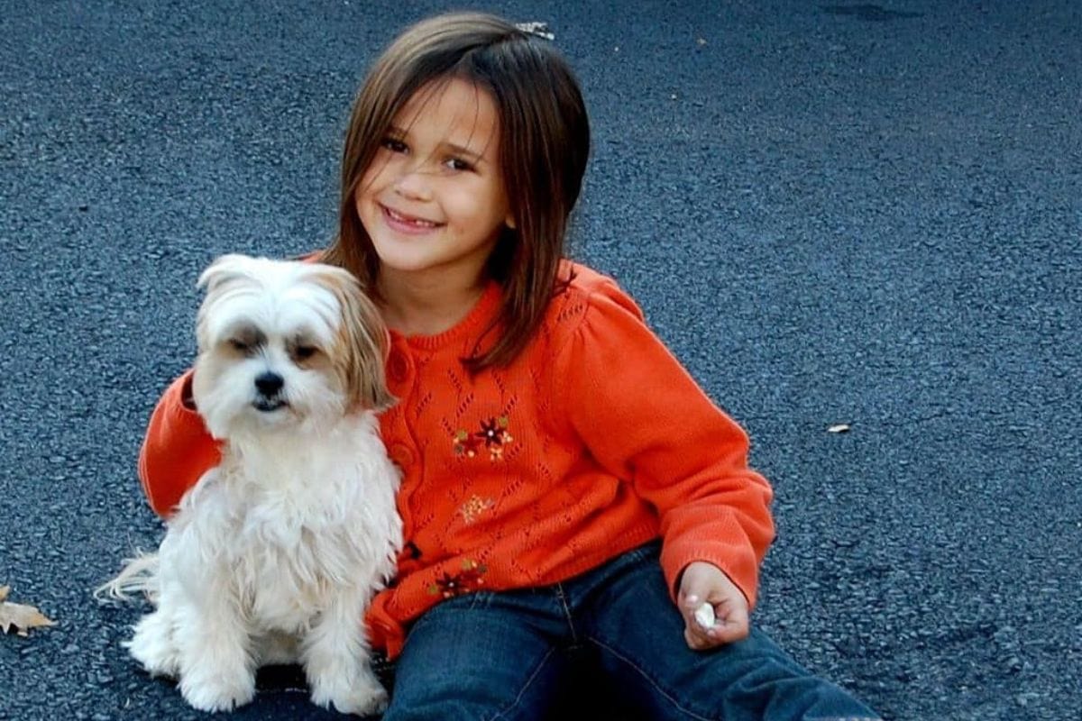 A young girl in an orange sweater sits on pavement, smiling, with her arm around a small white dog.