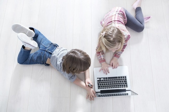 a boy and girl laying on a white wood floor looking at a laptop