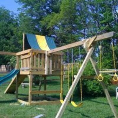 Wooden backyard playset featuring two swings, a slide, sandbox, climbing wall, and a green and yellow canopy—set on grass with trees behind. This backyard play equipment is worth the investment for endless outdoor fun.